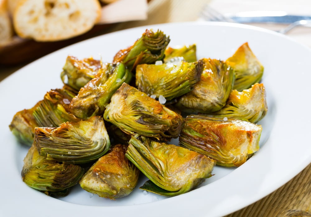 Fried artichoke hearts served on a white plate with coarse salt and bread in the background.