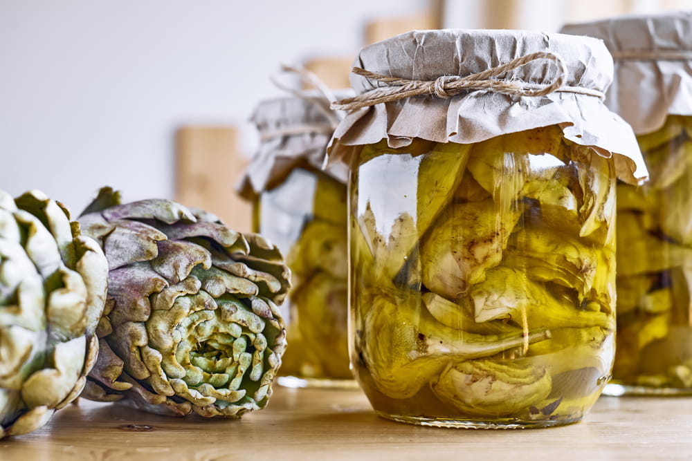 Jars of preserved artichokes in olive oil alongside fresh artichokes, a traditional Mediterranean way of preserving seasonal produce.